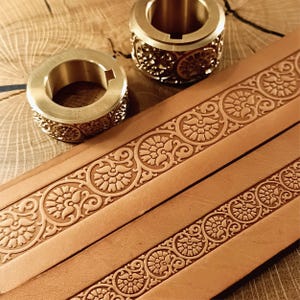 Puede incluir: Primer plano de dos anillos de latón con motivos florales y una tira de cuero marrón con diseños florales en relieve. Los anillos tienen un acabado metálico brillante. La tira de cuero muestra patrones florales simétricos detallados.