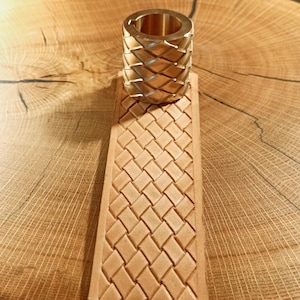 Puede incluir: Un cilindro de latón con un patrón tejido se asienta sobre una correa de cuero marrón claro con un diseño tejido a juego. Los artículos se exhiben sobre una superficie de madera con vetas naturales.