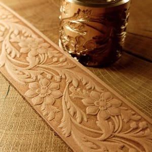 Puede incluir: Primer plano de un cinturón de cuero marrón claro y un objeto cilíndrico, ambos con intrincados grabados florales. El cinturón muestra un patrón repetitivo de flores y hojas. El objeto cilíndrico tiene una parte superior metálica.