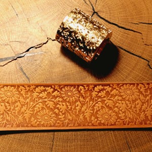 May include: A cylindrical gold-toned metal stamping tool featuring a detailed floral pattern with daisy-like flowers and leaves, positioned above a matching tan leather strip embossed with the same repeating floral design across its entire surface.