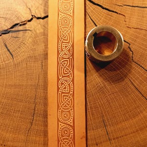 Puede incluir: Primer plano de una tira de cuero marrón claro con un diseño de nudo celta en relieve. Un anillo de metal de latón está sobre una superficie de madera. La tira de cuero es vertical.