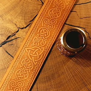 May include: A tan leather belt featuring an embossed pattern of intricate Celtic knots, spirals, and stylized animal figures, accompanied by a cylindrical metallic object with a textured surface and a smooth, circular top opening.