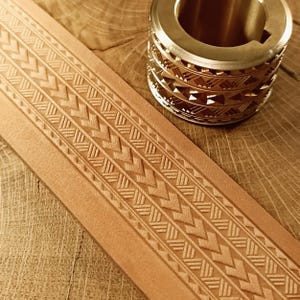 May include: A close-up shot of a tan leather strip with an embossed geometric pattern. A brass leather stamping tool is in the background. The leather strip and tool are on a wooden surface.