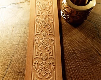 May include: A light brown leather strip with an embossed geometric pattern and a brass cylinder with a decorative design. The leather strip is rectangular and features a repeating symmetrical design. The brass cylinder has an open centre and an intricate pattern.
