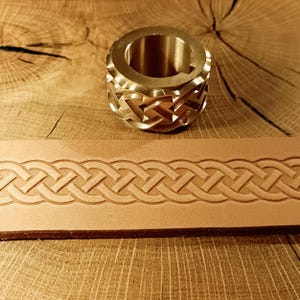 Puede incluir: Una herramienta de metal de latón con un diseño de nudo celta, junto a una tira de cuero marrón claro grabada con un patrón similar. La herramienta tiene forma cilíndrica con un centro hueco. La tira de cuero muestra un diseño tejido repetitivo.