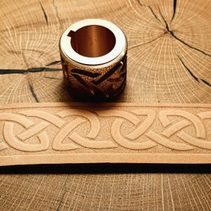 Op de afbeelding: Een cilindrisch, goudkleurig metalen object met een decoratief Keltisch knoopontwerp. Een lichtbruine leren riem met een Keltisch knoopmotief is ook zichtbaar. De items worden op een houten oppervlak weergegeven.
