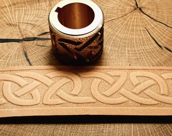 May include: A cylindrical, gold-colored metal object with a decorative Celtic knot design. A light brown leather belt with a Celtic knot pattern is also visible. The items are displayed on a wooden surface.