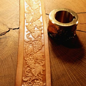 Pode incluir: Uma tira de couro bege com um padrão em relevo contínuo de cachos de uva e folhas de videira, posicionada ao lado de uma ferramenta cilíndrica de latão com um design gravado correspondente de uvas e vinhas na sua superfície externa.