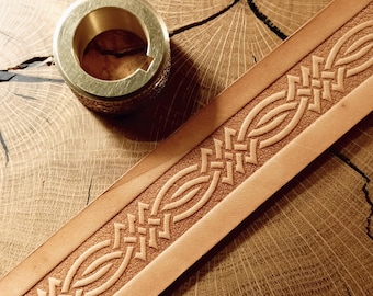 May include: A close-up of a light brown leather belt with an embossed geometric pattern. A brass metal ring sits next to the belt on a wooden surface. The belt has a detailed, textured design.