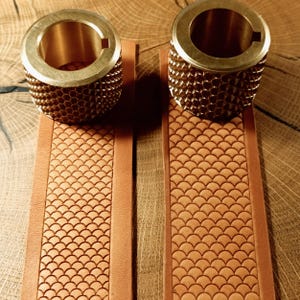 Puede incluir: Dos cilindros de latón con superficies texturizadas se asientan sobre dos correas de cuero marrón con un patrón de escamas. Los cilindros tienen un centro hueco y un borde liso de color dorado. Las correas de cuero son de color marrón claro.