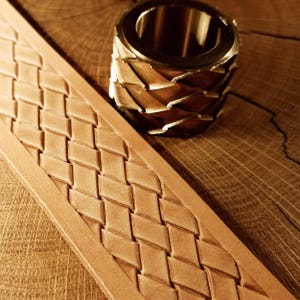 Puede incluir: Primer plano de una tira de cuero marrón claro con un patrón tejido, junto a una herramienta de metal de latón. La tira de cuero es de color marrón claro, con un diseño tejido en forma de diamante. La herramienta de latón es cilíndrica con un patrón decorativo.