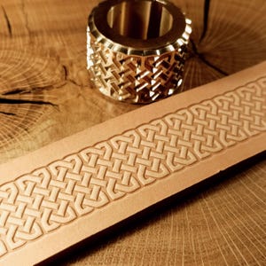 May include: A brass leather stamping tool and a strip of stamped leather. The tool is cylindrical with a geometric pattern. The leather strip displays a repeating Celtic knot design, embossed into the light brown leather.