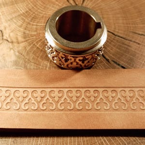 Puede incluir: Una herramienta de estampado de cuero de latón con un diseño decorativo. La herramienta se utiliza para crear un borde en artículos de cuero.