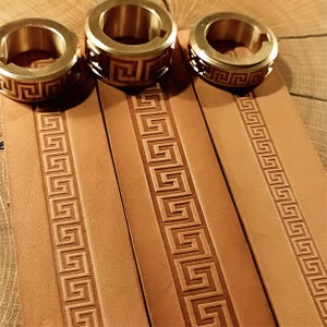 Op de afbeelding: Drie goudkleurige ringen met een geometrisch ontwerp liggen boven drie bruinleren riemen, elk met een reliëfpatroon. De ringen hebben een cirkelvorm met een gedetailleerd, opengewerkt ontwerp. De leren riemen zijn verticaal geplaatst.