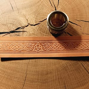 Puede incluir: Un cinturón de cuero marrón claro con un diseño de nudo celta y un anillo de latón. El cinturón presenta intrincados patrones en relieve de líneas entrelazadas y figuras de animales estilizadas. El anillo de latón tiene una superficie texturizada.