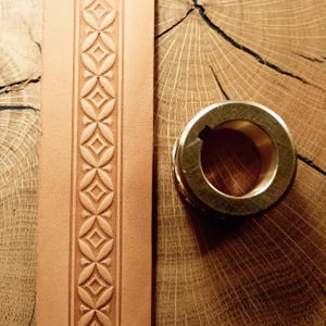 Puede incluir: Primer plano de un cinturón de cuero color beige con un patrón geométrico repetido. Junto a él, un anillo metálico dorado con una ranura para chaveta. Los objetos están sobre una superficie de madera.