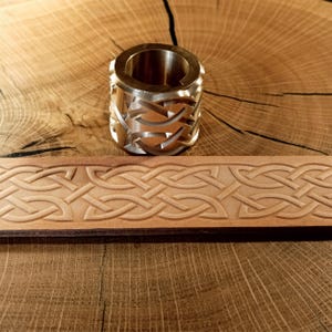 Puede incluir: Una cuenta cilíndrica de metal plateado con un diseño de nudo celta, junto a una tira de cuero marrón claro con un patrón en relieve a juego. La cuenta tiene secciones recortadas. La tira de cuero es de un tono más claro.