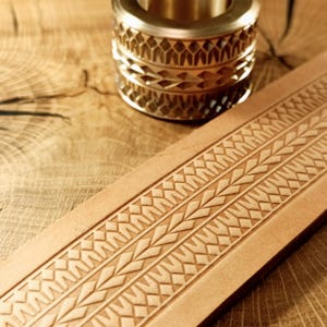 May include: A close-up of a gold-colored ring with a geometric pattern and a light brown leather strap with an embossed design. The ring is cylindrical with multiple bands of detailed carvings. The leather strap has a repeating pattern of shapes.