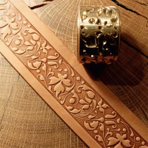 May include: A tan leather belt with an embossed floral pattern and a gold-coloured metal stamp. The stamp has a matching floral design. The belt and stamp are on a wooden surface, showing the tooling process.