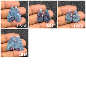 以下が含まれることがあります： 4組のしずく型の宝石。石は青灰色のベースカラーで、赤とピンクのインクルージョンがあります。各ペアは白いテキストでラベル付けされています：L613、L614、L615、およびL616。宝石は手の上に表示されています。
