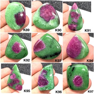 Può includere: Collezione di pietre preziose lucide con una vibrante matrice verde e inclusioni rosso scuro. Le pietre variano nella forma, tra cui ovale, triangolare e a goccia. Ogni pietra ha un motivo unico ed è etichettata con una 'K' seguita da un numero.