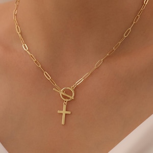 May include: A gold-colored necklace with a cross pendant. The necklace features a toggle clasp and a chain with oval links. The cross pendant hangs from a circular bail. The necklace is displayed against a white background.