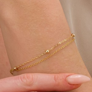 Puede incluir: Una delicada pulsera dorada con dos cadenas paralelas y pequeñas cuentas espaciadas. La pulsera se lleva en la muñeca, mostrando su elegancia sencilla y brillo sutil. La pieza de joyería es un accesorio clásico.