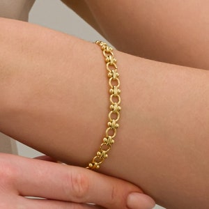 May include: A gold bracelet featuring a repeating pattern of linked circles and small, round details. The bracelet is displayed on a wrist, highlighting its design and colour. The jewellery item is a classic accessory.
