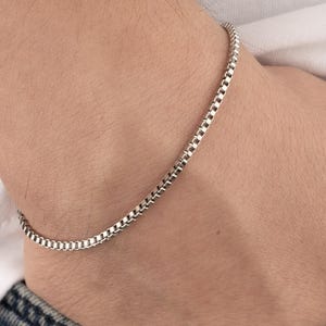 May include: A silver box chain bracelet. The bracelet is worn on a wrist, with the hand resting in a blue denim jeans pocket. The person is wearing a white t-shirt.