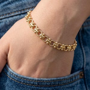 Op de afbeelding: Een gouden kettingarmband met een herhalend patroon van cirkelvormige schakels verbonden door kleine, gepolijste gouden bolletjes. Het armband is gemaakt van een reeks onderling verbonden ronde metalen lussen, wat zorgt voor een textuurrijk ontwerp.