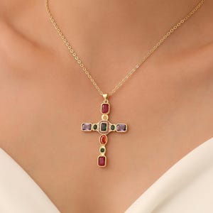 May include: A gold cross pendant necklace with a fine chain. The cross features an array of colourful gemstones, including red, purple, green, and black. The necklace is displayed against a white top.