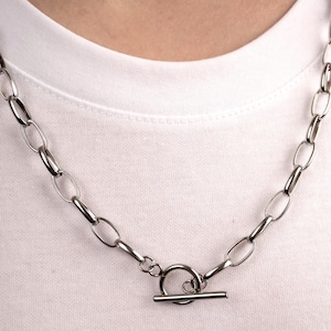 May include: A silver-colored chain necklace with an oval link design and a toggle clasp. The necklace is worn against a white t-shirt, creating a contrast. The jewelry is a simple, yet stylish accessory.