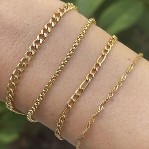 May include: Four gold chain bracelets of different designs are shown on a wrist. The bracelets include a curb chain, a beaded chain, a Figaro chain, and a twisted rope chain. The bracelets are all a shiny gold colour.