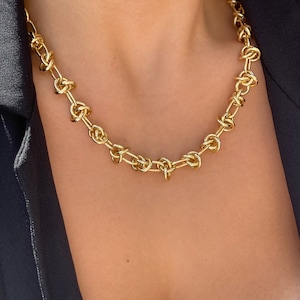 May include: A gold chain necklace with a knotted design. The necklace is worn by a person with a white top and a black blazer.