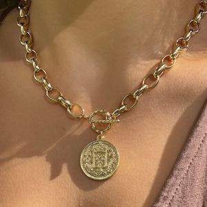 May include: A gold chain necklace with a large, ornate coin pendant. The chain is thick and has a chunky, oval link design. The pendant is round and features a detailed design.