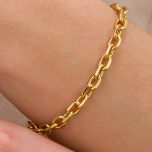 May include: A gold chain bracelet with oval links is shown on a person's arm. The bracelet has a bright, shiny gold colour and appears to be made of metal. The bracelet is worn on the wrist and is the main focus of the image.
