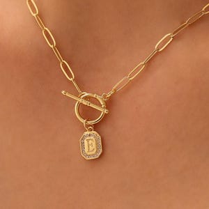 May include: A gold necklace featuring a toggle clasp and a rectangular pendant. The pendant displays the letter "E" surrounded by small, sparkling details. The chain is made of elongated oval links.