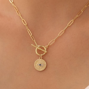 May include: A gold-colored necklace with a toggle clasp and a circular pendant. The pendant features an eye design with a small blue stone in the center. The chain has an oval link design.