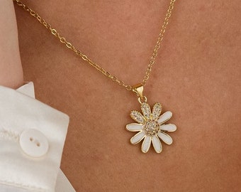 May include: A gold-colored daisy pendant necklace with a white enamel center and clear crystal accents. The necklace is on a gold chain. The person is wearing a white button-down shirt.