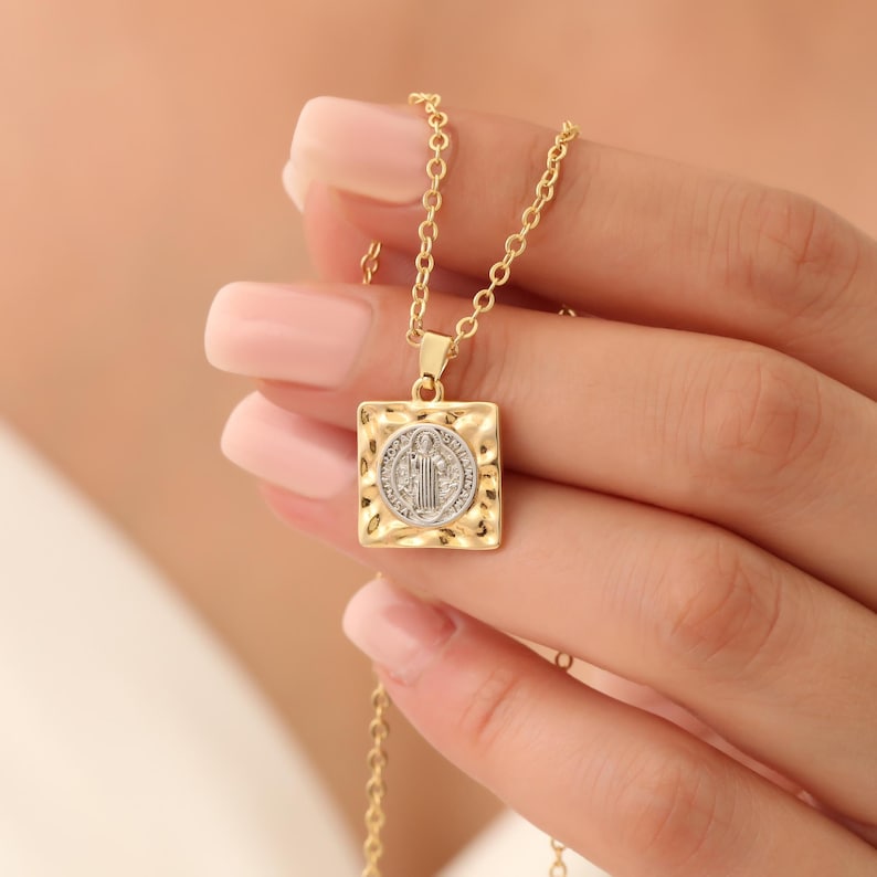 May include: Gold chain necklace with a square pendant featuring a Saint Benedict medal. The pendant has a textured, hammered finish.