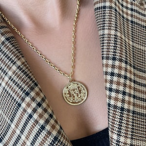 May include: A gold chain necklace with a large, round pendant featuring a detailed design. The pendant has a crown and other intricate details.