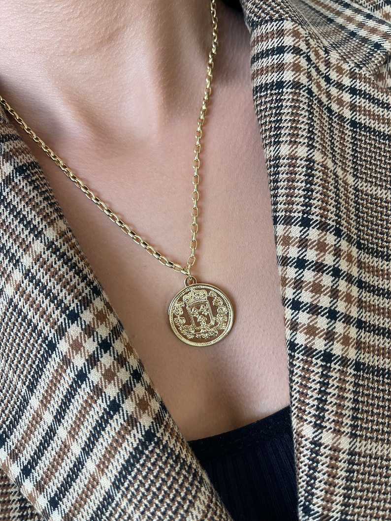 May include: A gold chain necklace with a large, round pendant featuring a detailed design. The pendant has a crown and other intricate details.