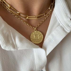 May include: A gold chain necklace with a large, round, gold coin pendant. The coin has intricate details and a raised design.