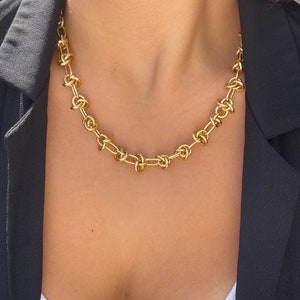 Peut inclure: Un collier en cha&icirc;ne dor&eacute;e avec un motif nou&eacute;. Le collier est port&eacute; par une personne avec un blazer noir et une chemise blanche.