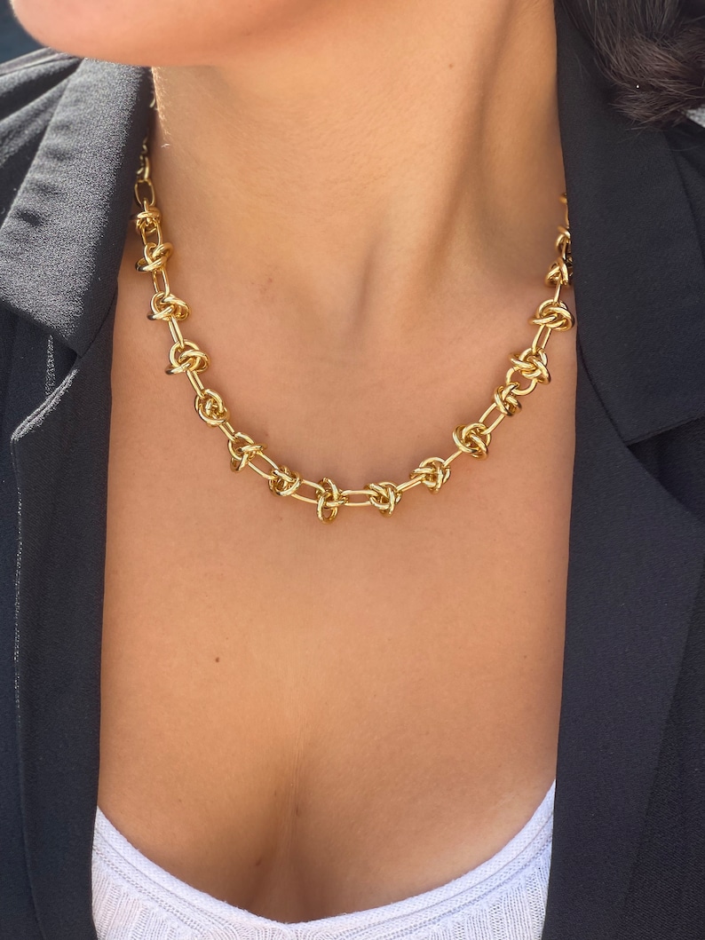 Peut inclure: Un collier en cha&icirc;ne dor&eacute;e avec un motif nou&eacute;. Le collier est port&eacute; par une personne avec un blazer noir et une chemise blanche.