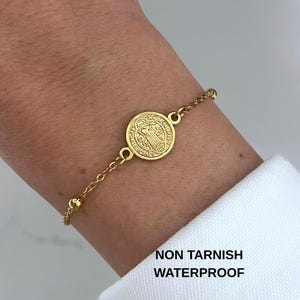 May include: Gold-colored bracelet featuring a round pendant with a religious figure. The bracelet has a delicate chain design with small ball accents. The text "NON TARNISH WATERPROOF" is visible in the image.