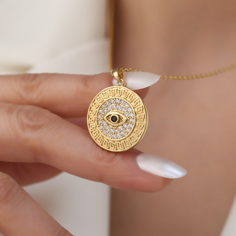 May include: Gold necklace with a round pendant featuring a black evil eye surrounded by clear cubic zirconia stones. The pendant is decorated with a Greek key pattern.