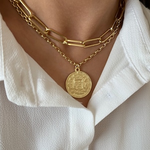 May include: A gold chain necklace with a large, round pendant featuring a detailed design. The pendant is layered over a second, thicker gold chain.