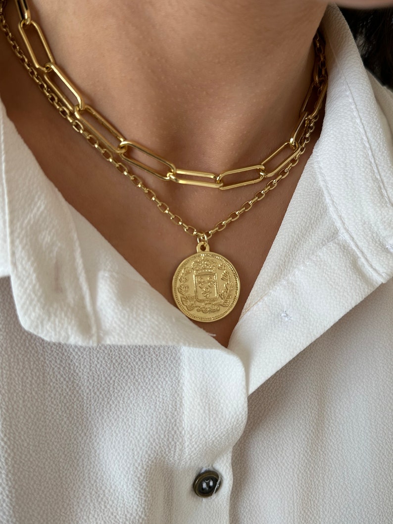 May include: A gold chain necklace with a large, round pendant featuring a detailed design. The pendant is layered over a second, thicker gold chain.
