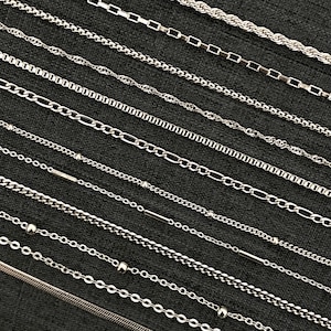 Puede incluir: Una selección de collares plateados presentados sobre un fondo gris oscuro. Las cadenas presentan diversos diseños, como estilos de cuerda, caja, cable y cuentas. Los collares están dispuestos en diagonal, destacando sus variadas texturas y patrones.